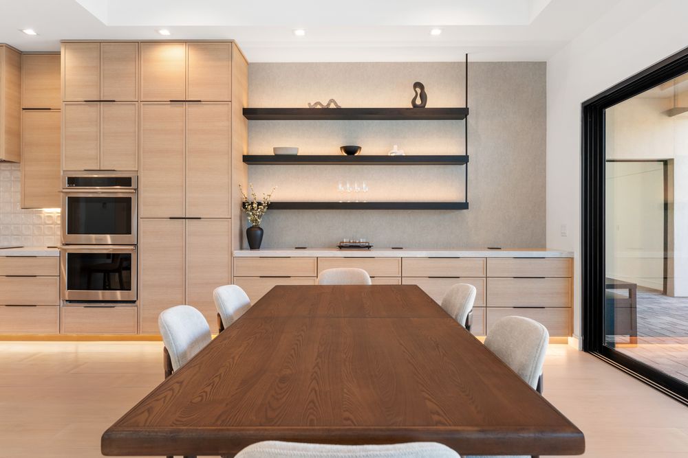 Dining table in front of the double ovens and lit floating shelves